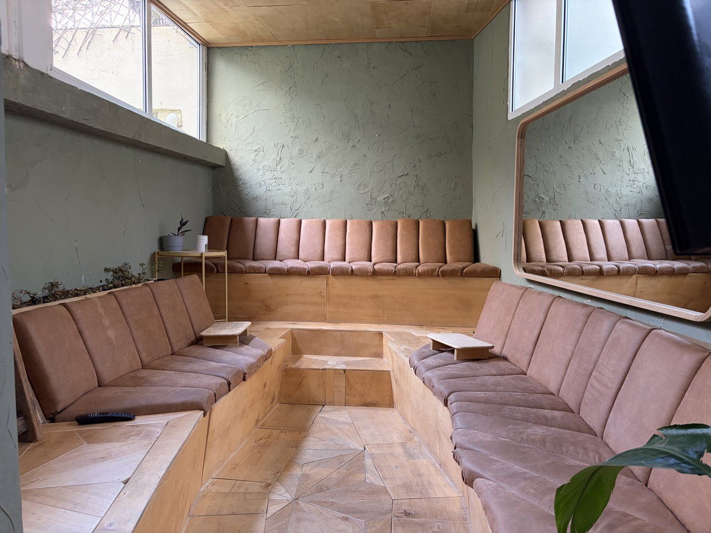 The Amphitheatre: a horseshoe of tan velvet banquette seating wrapped around low wooden tables, sage plaster walls, and daylight from the windows above the seats.