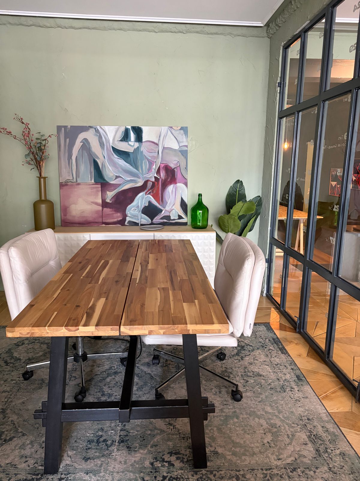 The Salon: a second wooden table set with chairs, framed by iron-framed industrial glazing, with artwork on the sage wall.