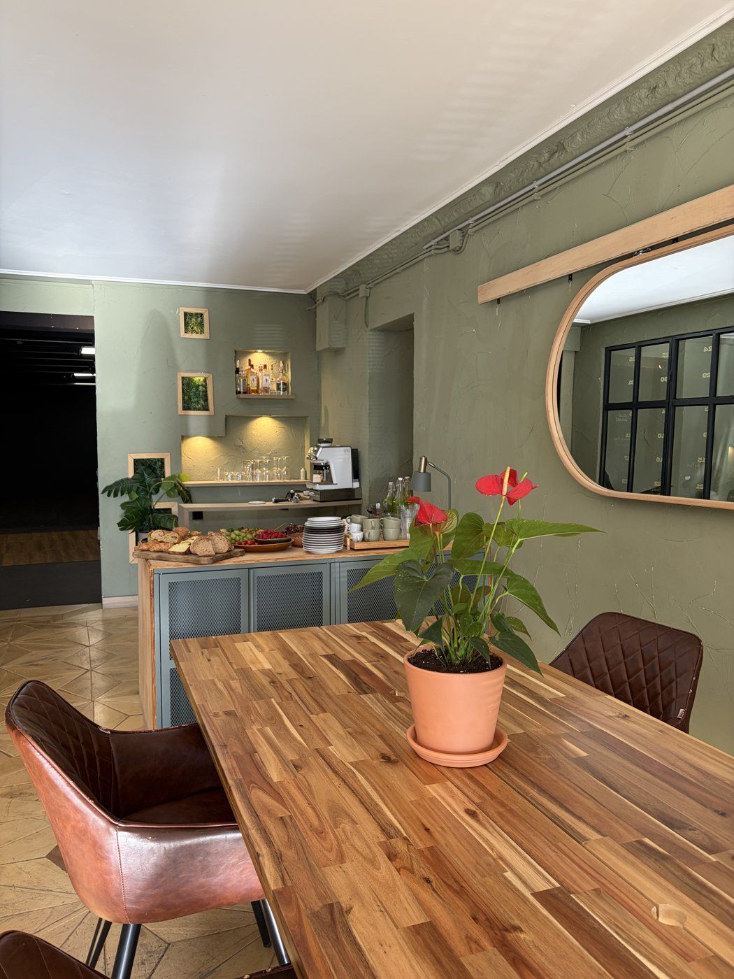 The Forum: a long wooden table with leather chairs, the drinks and coffee bench behind it, sage plaster walls and an arched mirror.